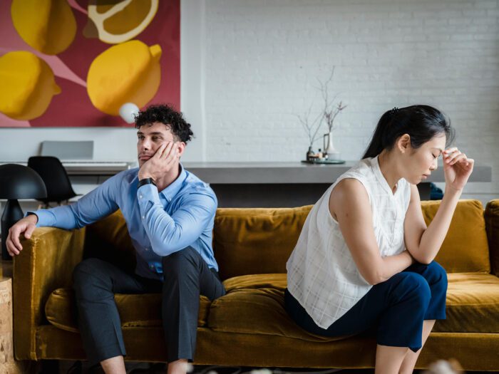 Couple sitting back to back on a sofa looking upset